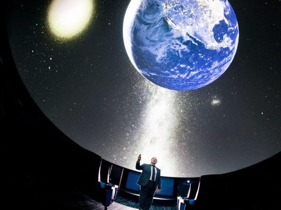 Image of a planetarium with a model of earth suspended in the air and a man standing under it pointing towards it.